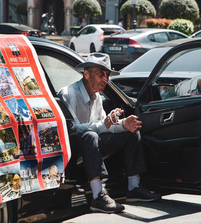 Senior man in hat sits in car displaying a travel poster. Urban street scene.